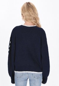 Marineblauer Pullover mit einem flauschigen Textur, überschnittenen Schultern und weißen Akzenten entlang des Ausschnitts und der Ärmel. Kombiniert mit hellblauen Jeans.