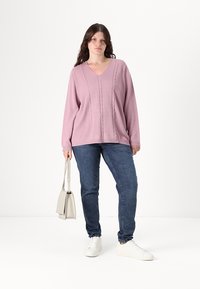 Ανοιχτό μωβ πλεκτό πουλόβερ με V-ντεκολτέ και σχέδιο καμπάνας, σε συνδυασμό με σκούρα μπλε τζιν και λευκά αθλητικά παπούτσια. Κρατάει μια μικρή μπεζ τσάντα.