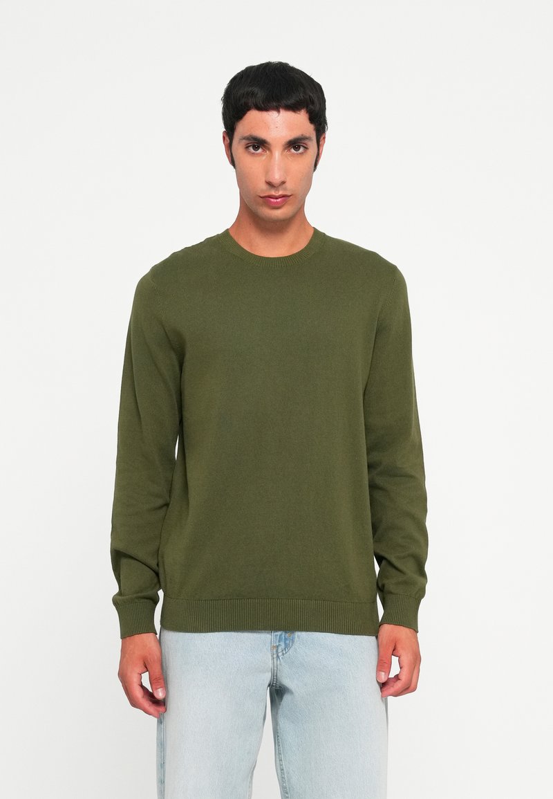 Groene gebreide trui met een ronde hals en lange mouwen, voorzien van ribgebreide boorden en onderrand, gecombineerd met lichtblauwe jeans.