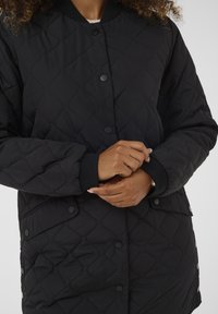 Person passt schwarze gesteppte Jacke mit Druckknöpfen und gerippten Bündchen an, zeigt oberen Rumpf und Hände vor einfarbigem Hintergrund.