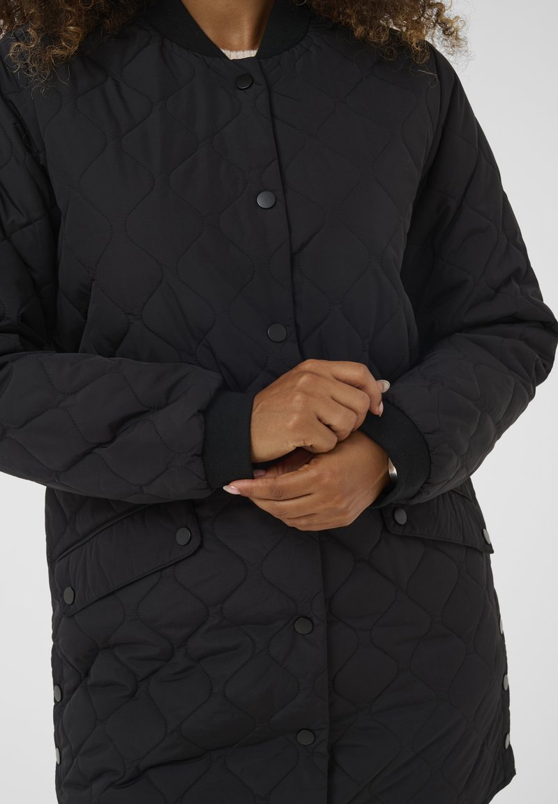 Person passt schwarze gesteppte Jacke mit Druckknöpfen und gerippten Bündchen an, zeigt oberen Rumpf und Hände vor einfarbigem Hintergrund.