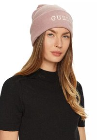 Bonnet côtelé rose avec logo brodé "GUESS". Porté avec un haut noir à manches courtes. Texture douce et coupe décontractée.