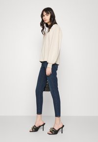 Beige dlouhý rukáv top s nabíranými rukávy, tmavě modré skinny džíny a černé sandály na podpatku s otevřenou špičkou a zlatými detaily.
