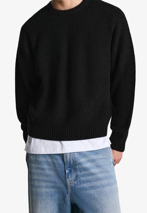 Homme portant un pull en maille côtelée noire sur une chemise blanche et un jean bleu clair sur fond blanc.