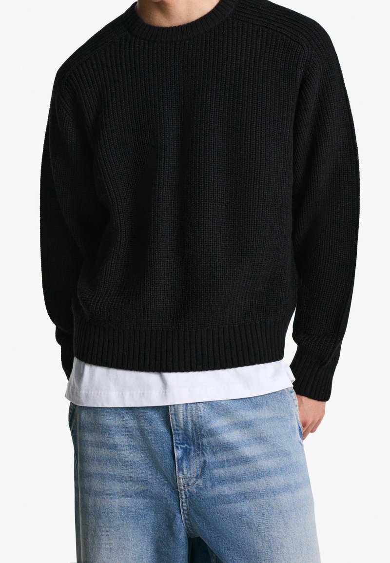 Homme portant un pull en maille côtelée noire sur une chemise blanche et un jean bleu clair sur fond blanc.