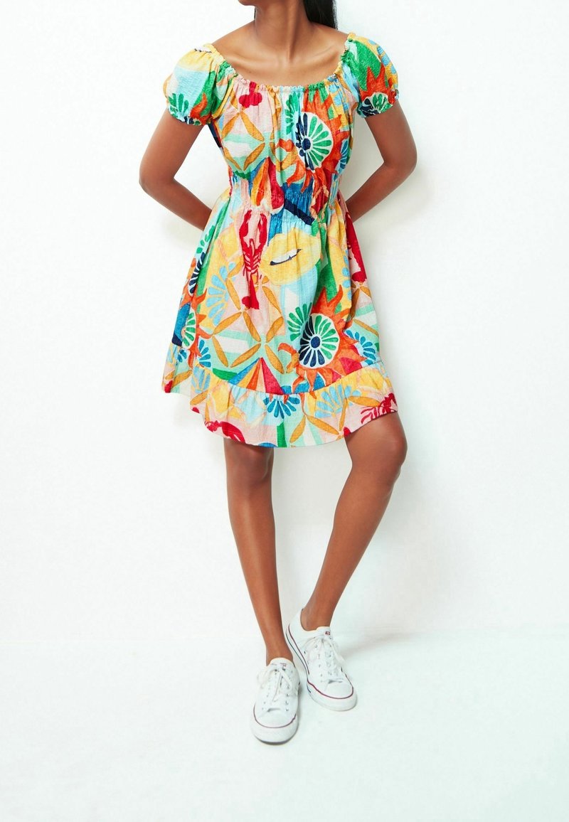 Femme portant une robe courte colorée à motifs floraux avec des manches bouffantes et des baskets blanches, debout les mains derrière le dos contre un mur blanc.