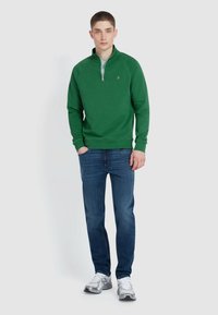 Green zip-neck sweatshirt with ribbed cuffs, paired with blue denim jeans and gray sneakers. Solid colors, casual fit, and simple design.