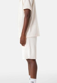 Beige top met korte mouwen en textuur, gecombineerd met bijpassende shorts, beide met een subtiel geribbeld patroon. Effen witte sokken met zwarte tekst.