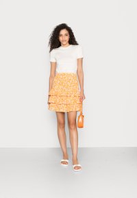 Top ajustado de mangas curtas brancas, saia amarela com camadas e babados de flores, sandálias brancas e uma bolsa transversal laranja. Paleta de cores simples e vibrante.