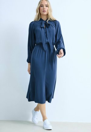 Vrouw loopt en draagt een marineblauwe midi-jurk met lange mouwen, een neklint en witte sneakers, tegen een effen lichte achtergrond.