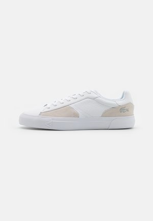 Baskets blanches basses avec des accents en daim beige, des lacets blancs et un petit logo crocodile brodé près du talon sur une semelle blanche.