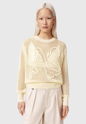 Junge Frau mit langen blonden Haaren, die einen beigen Mesh-Pullover mit großen bestickten Buchstaben und hellfarbene plissierte Hosen trägt.