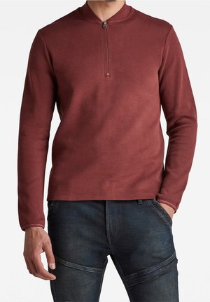 Chemise à manches longues bordeaux avec un col montant et une demi-fermeture éclair. Fabriquée dans un matériau doux, avec une texture unie et une silhouette ajustée. Associée à un jean bleu.