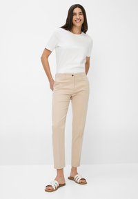 Beige op maat gemaakte broek met een slim fit, voorzakken en een enkele knoopsluiting. Gecombineerd met een eenvoudig wit T-shirt met korte mouwen en witte sandalen.
