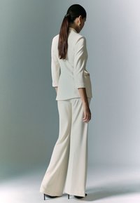 Completo su misura di un beige chiaro con giacca aderente e pantaloni a gamba larga, caratterizzato da una texture liscia e cuciture minime, abbinato a tacchi neri.