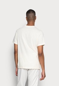 Kremowa bawełniana koszulka z klasycznym okrągłym dekoltem, krótkimi rękawami i luźnym krojem, widziana z tyłu, bez widocznych logotypów ani wzorów.