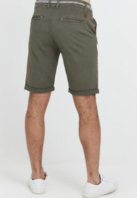 Shorts olive green avec revers en tissu lisse, dotés de poches arrière et d'une ceinture à motif. Portés avec des baskets blanches.