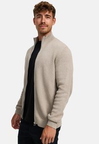 Beige ritstrui met textuurpatroon, geribde manchetten en kraag, gedragen over een zwart shirt. Effen achtergrond.