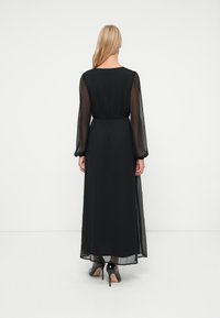 Frau mit langen blonden Haaren trägt ein schwarzes durchsichtiges Maxikleid mit langen Ärmeln und schwarze High Heels und steht vor einer weißen Wand.