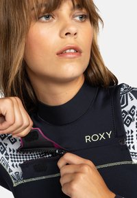 Zwarte wetsuit met gedessineerde mouwen, Roxy-logo, roze accenten en een ritssluiting. Heeft een hoge kraag en textuurstof.