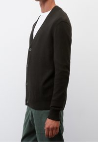 Man som bär en svart knappad cardigan över en vit skjorta och gröna byxor, avbildad från axlarna till mitten av låren mot en enkel bakgrund.