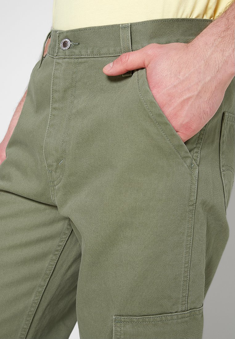 Pantalons cargo en coton vert olive avec une coupe droite, dotés de poches latérales et d'une fermeture à bouton, montrés avec une main dans la poche avant.