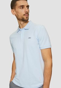 Mann in hellblauem s.Oliver Polohemd mit kurzen Ärmeln, stehend mit Händen in den Taschen und zur Seite schauend.