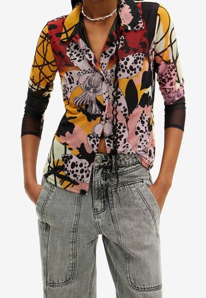 Woman wearing a colorful floral patterned shirt over a sheer black long sleeve top, paired with high-waisted gray denim pants.