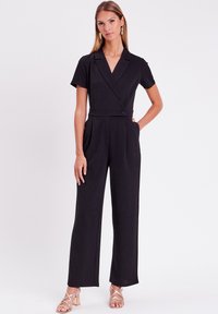 Une combinaison noire à manches courtes, avec un devant croisé, un détail bouton à la taille, et un bas à jambes larges, associée à des talons à lanières.