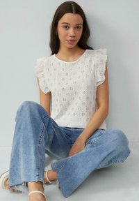 Weiße Bluse mit Lochmuster und Rüsche Ärmeln, kombiniert mit hellblauen, weiten Jeans und weißen Sandalen. Glatte Textur und lässiges Design.