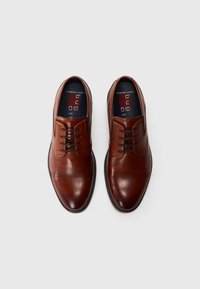 Chaussures de cérémonie en cuir marron avec une finition brillante, présentant un design à lacets, des coutures décoratives et un ajustement large confortable.