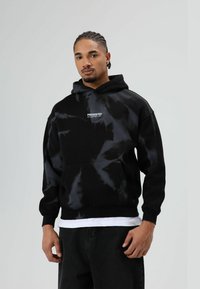 Zwarte tie-dye hoodie gemaakt van katoen. Voorzien van een voorzak, geribbelde manchetten en een gedrukt logo op de borst. Model poseert tegen een neutrale achtergrond.
