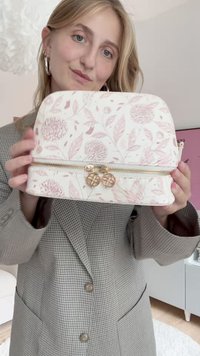 Weiße Tasche mit pinkfarbigem Blumenmuster, rundem Oberteil, goldfarbenem Reißverschluss und doppelten Charms. Aus strukturiertem Stoff gefertigt und mit zarten Blattmustern versehen.