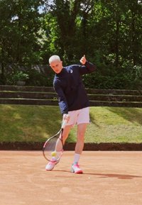 Un joueur de tennis masculin frappe une balle sur un court en terre battue. Il porte une chemise à manches longues bleu et un short blanc, avec des chaussures à accents roses.