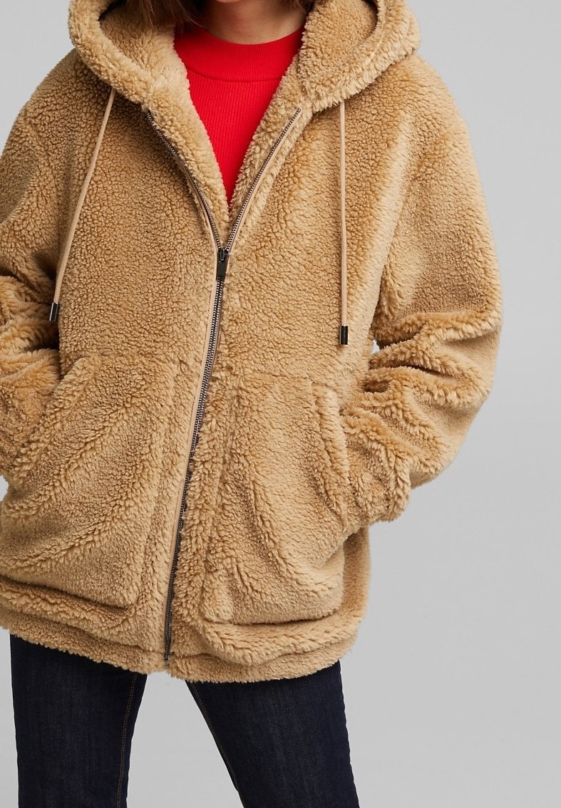 Personne portant une veste à capuche beige duveteuse avec fermeture éclair sur un t-shirt rouge et un jean foncé, les mains dans les poches avant.