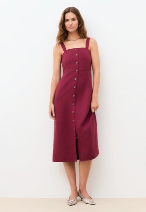 Frau in einem ärmellosen, burgunderfarbenen Midikleid mit Knopfleiste vorne und quadratischem Ausschnitt, trägt schlangenprintabsatzschuhe und steht vor einer weißen Wand.