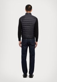 Gilet matelassé noir avec des coutures horizontales, associé à une chemise à manches longues noire texturée et un jean bleu foncé. Posture debout, de dos.