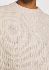 Maglione lavorato a maglia di un beige chiaro con scollo a girocollo a coste, caratterizzato da una morbida texture e da un motivo di sottili linee verticali.