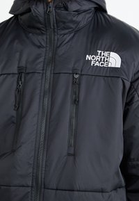 Černá zateplená bunda s kapucí, obsahující zipové kapsy a bílé logo na levé hrudi. Hladká textura a lehký design.