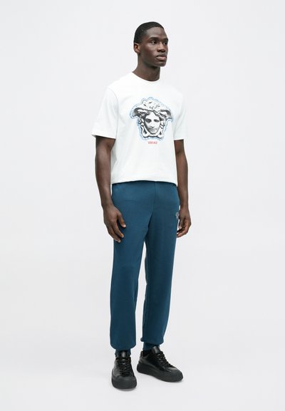 Λευκό κοντομάνικο T-shirt με γραφικό μιας φιγούρας περιβαλλόμενης από περγαμηνές. Συνδυάζεται με σκούρο μπλε παντελόνι και μαύρα αθλητικά παπούτσια.