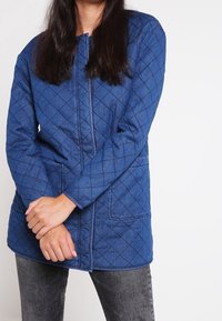 Blå quiltad jacka med dragkedja framtill, långa ärmar och två fickor. Har ett diamantmönster och en avslappnad passform, kombinerad med mörka jeans.