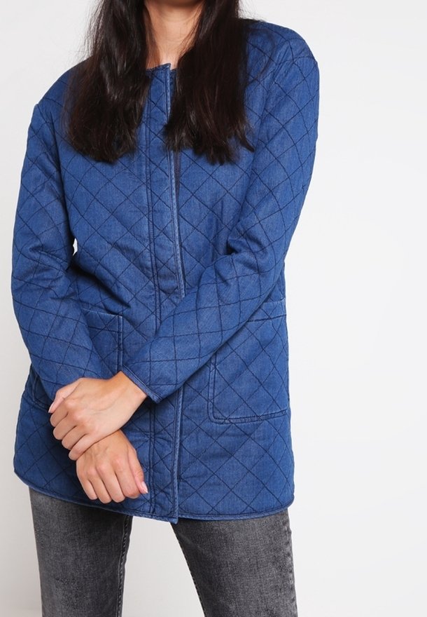 Blå quiltad jacka med dragkedja framtill, långa ärmar och två fickor. Har ett diamantmönster och en avslappnad passform, kombinerad med mörka jeans.