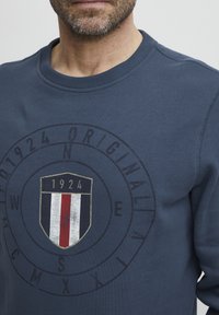 Marineblå hettegenser med et rundt grafisk motiv som har et skjolddesign og året "1924." Teksten rundt grafikken inkluderer retningbokstaver.