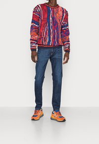 Man in een levendig, multicolor gebreid patroon trui, donkerblauwe jeans en feloranje en blauwe sneakers, staande op een effen witte vloer.