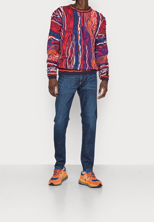 Homme portant un pull à motifs multicolores éclatants, un jean bleu foncé et des baskets orange et bleues vives, se tenant sur un sol blanc uni.
