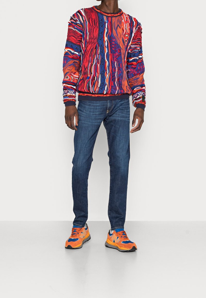 Man in een levendig, multicolor gebreid patroon trui, donkerblauwe jeans en feloranje en blauwe sneakers, staande op een effen witte vloer.