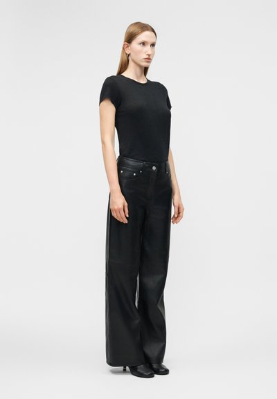 Γυναίκα που στέκεται, φορώντας μαύρη κοντομάνικη μπλούζα, μαύρο δερμάτινο παντελόνι με φαρδύ πόδι και μαύρα παπούτσια με τακούνι, σε απλό ανοιχτό γκρι φόντο.