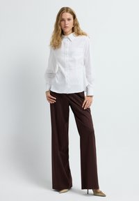Una donna sta in piedi davanti a uno sfondo bianco indossando una camicia bianca aderente con bottoni, pantaloni a righe marroni a gamba larga e decolletè beige a punta.