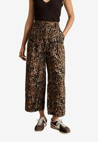 Pantalons à jambes larges en imprimé léopard de couleur brune et noire, avec une taille haute, des poches latérales et une coupe décontractée. Associés à des baskets sombres.