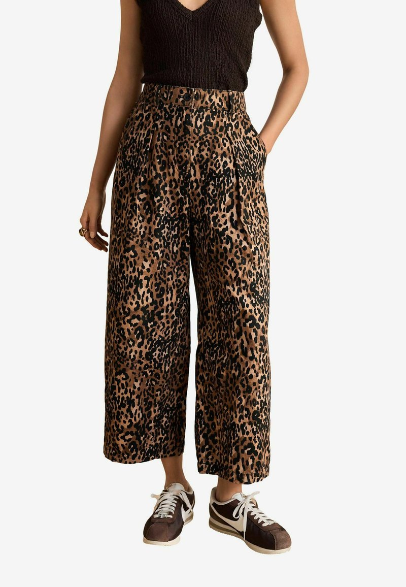 Pantalons à jambes larges en imprimé léopard de couleur brune et noire, avec une taille haute, des poches latérales et une coupe décontractée. Associés à des baskets sombres.